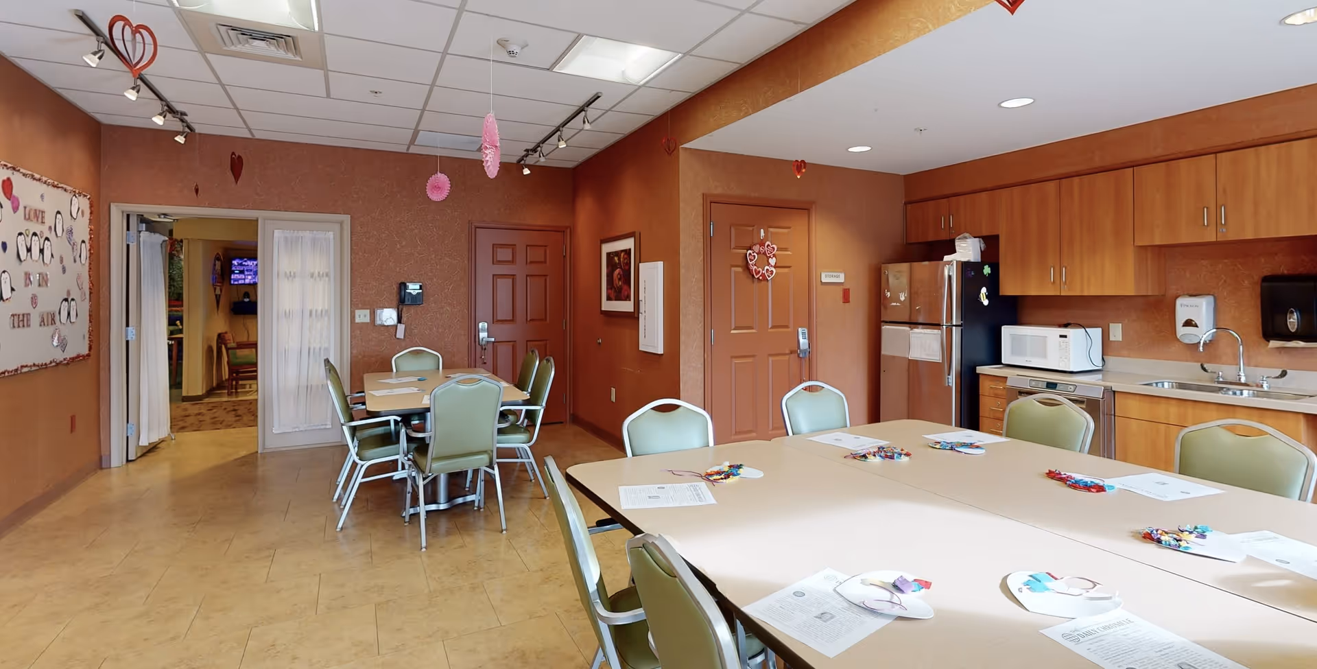 A communal dining and activity room with tables and green chairs arranged around them. The tables have papers and craft materials on them. The room has a kitchenette area with a refrigerator, microwave, dishwasher, sink, and wooden cabinets. The walls are decorated with heart-shaped hanging decorations and a bulletin board with penguin drawings and the words 'LOVE IS IN THE AIR'. There are two closed doors and an open doorway leading to another room with a TV and chairs.
