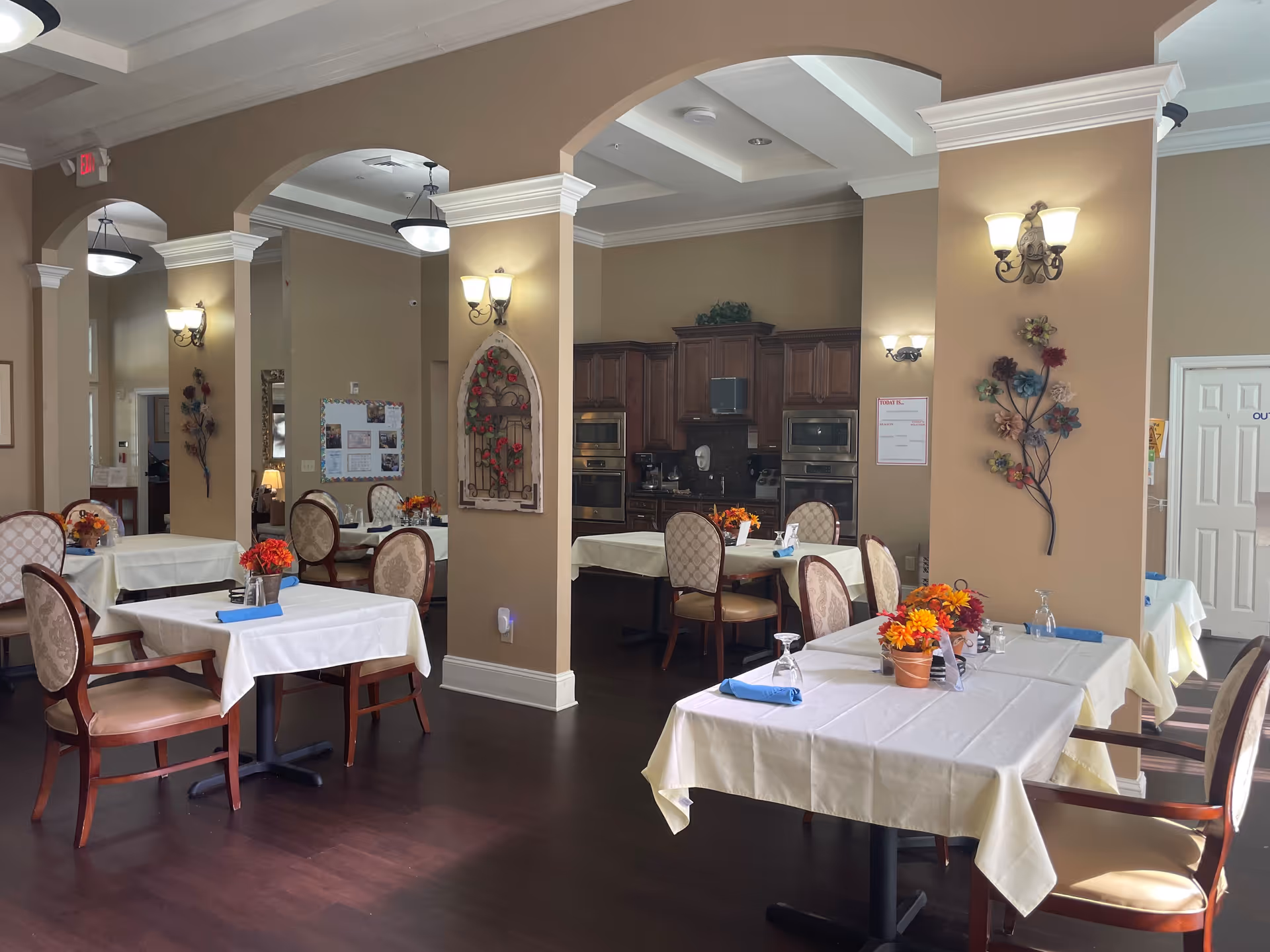A dining room in a senior living facility with several tables covered in white tablecloths, each set with blue napkins, glasses, and small flower arrangements. The room has beige walls, dark wood flooring, and decorative wall sconces. In the background, there is a kitchen area with wooden cabinets and stainless steel appliances.