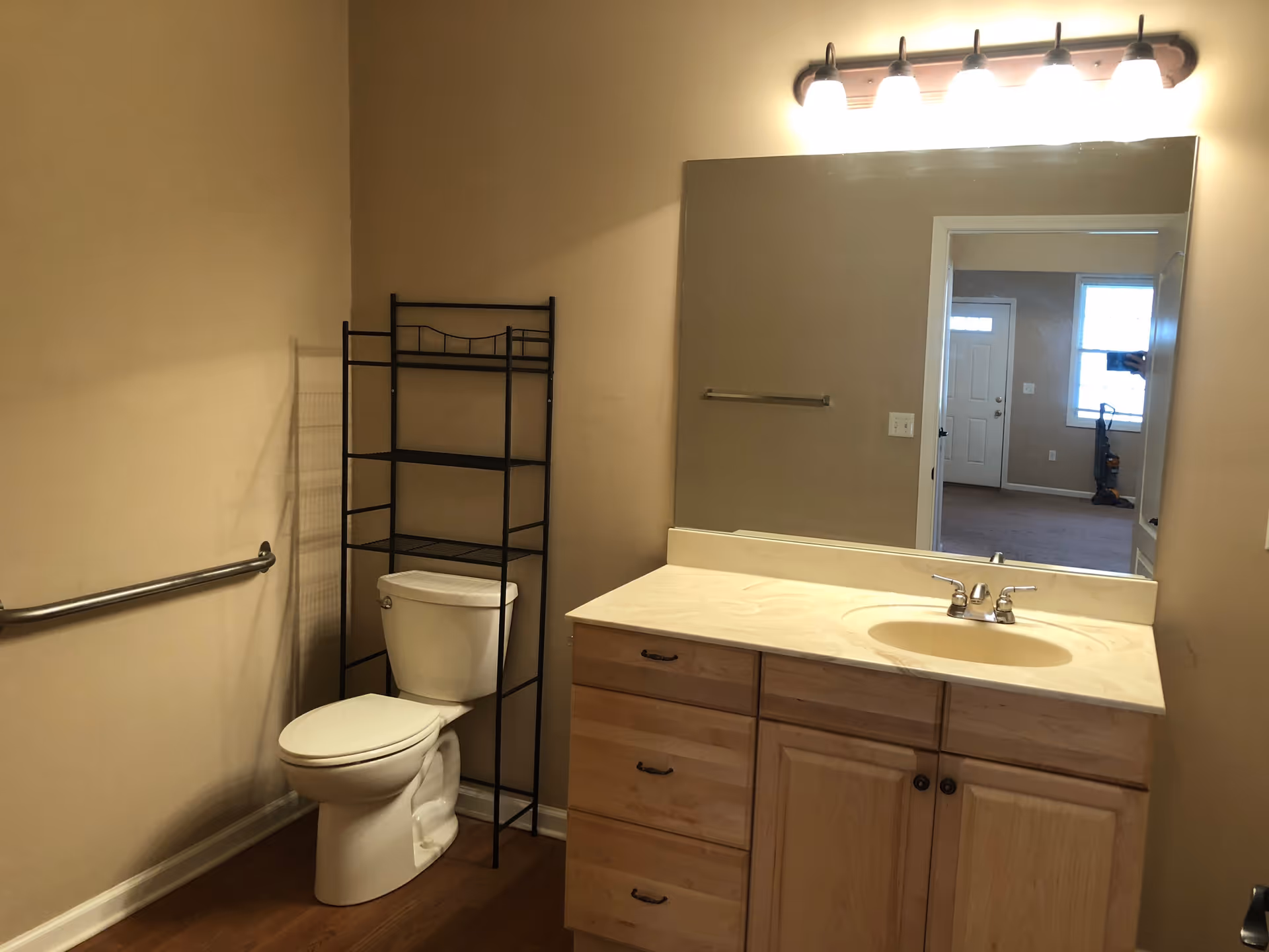 Bathroom showing a toilet with a black shelving unit, a wooden vanity with sink and large mirror, and a wall-mounted grab bar.