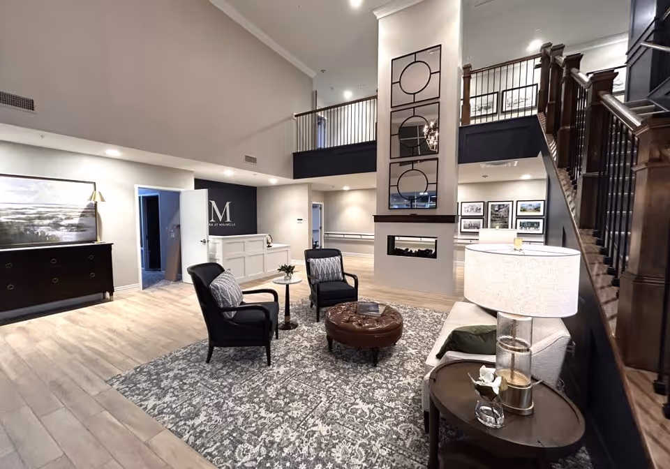 A spacious and modern senior living facility lobby with high ceilings, a patterned area rug, two black armchairs with cushions, a round brown ottoman, a white sofa with a side table and lamp, and a decorative fireplace with mirrors above it. There is a staircase with wooden railings on the right and a reception desk in the background with the letter M and the words 'Mira at Maumelle' on the wall behind it.