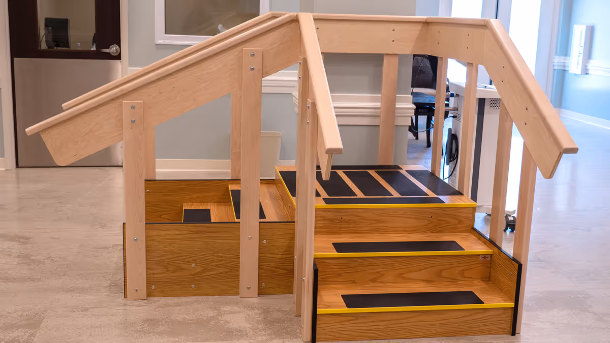 A wooden rehabilitation staircase with handrails on both sides and non-slip black strips on the steps, placed indoors on a light-colored floor with a door and windows in the background.
