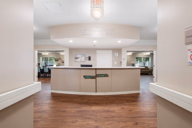 Reception desk area inside Home Place of New Bern facility with wooden flooring, beige walls, and a sign above the desk reading 'Home Place of New Bern'. There are two open doorways on either side of the desk leading to rooms with seating and windows.