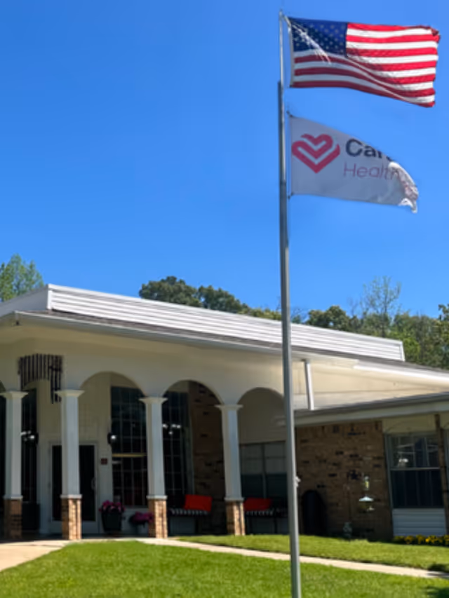 Exterior view of Caraday of Mineola facility showing a building with brick and white columns under a clear blue sky. Two flags are flying on a flagpole in front of the building, one is the American flag and the other is a Caraday Health flag. There is a green lawn and a walkway leading to the entrance.