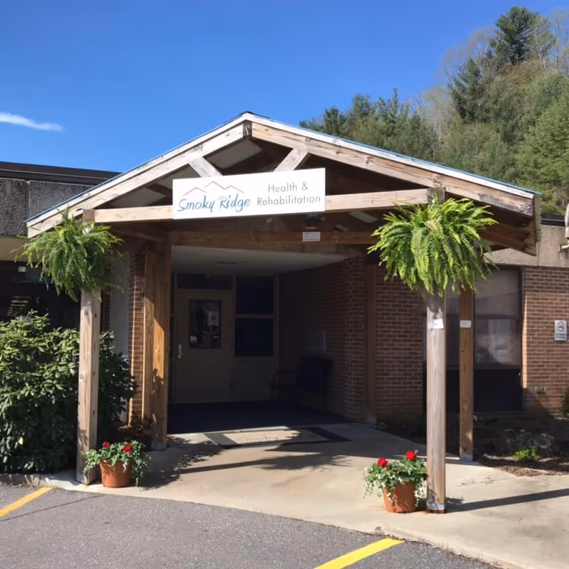 Entrance to Smoky Ridge Health & Rehabilitation facility with a wooden canopy supported by four posts, two hanging green ferns, and two potted plants with red flowers on either side of the walkway. The building is made of brick and there are trees visible in the background under a clear blue sky.