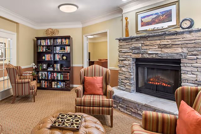 Cozy common room with a stone fireplace, striped armchairs, an ottoman, and a bookshelf.
