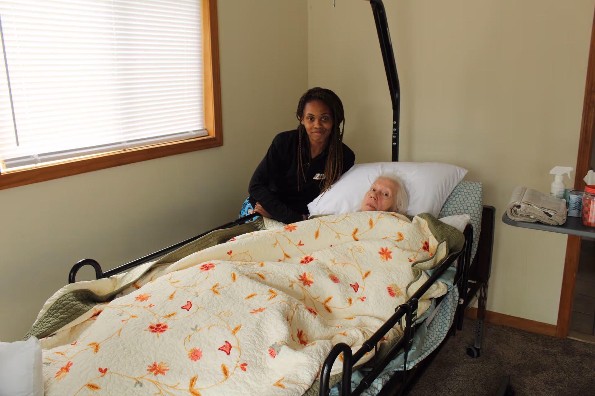 An elderly woman lying in a hospital-style bed covered with a floral quilt, with a caregiver sitting beside her. The room has beige walls, a window with closed blinds, and a small table with a newspaper, spray bottle, and other items.