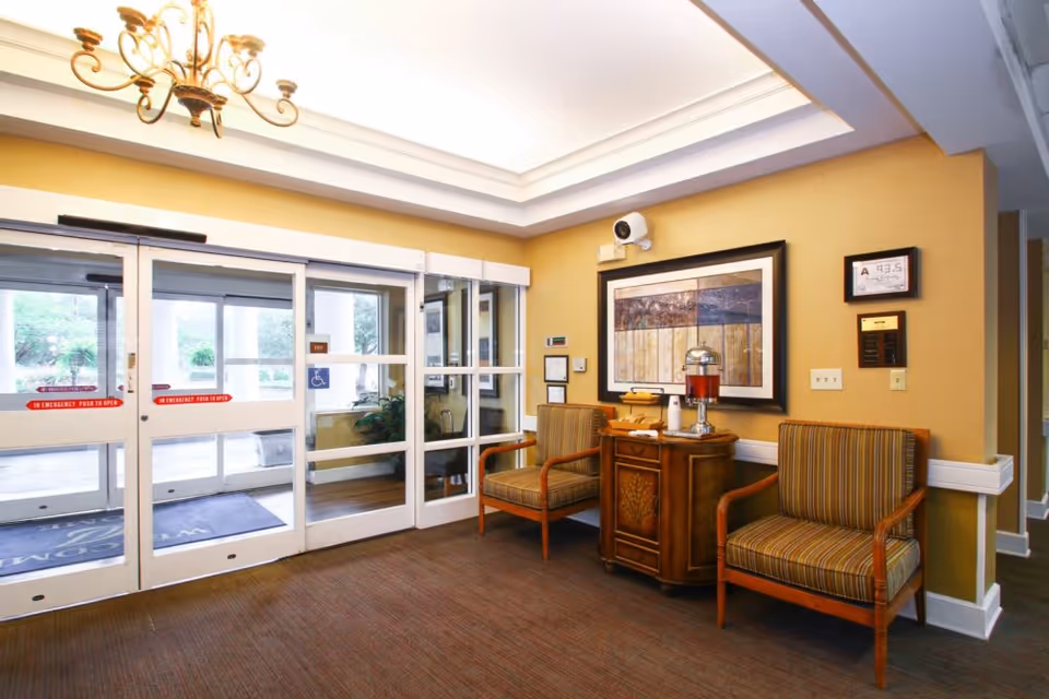 Entrance lobby with sliding glass doors, two striped upholstered chairs, a small wooden table holding a beverage dispenser, and framed artwork on the wall.