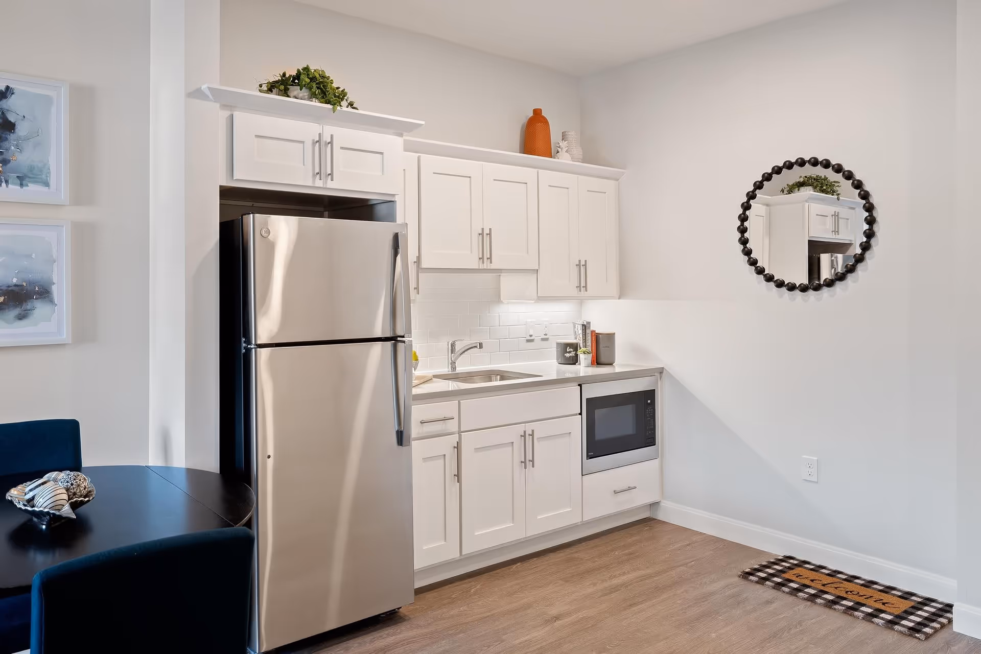 A modern kitchen area featuring white cabinets, a stainless steel refrigerator, a built-in microwave, and a sink with a faucet. There is a round mirror with a beaded frame on the wall, a small plant and decorative items on the countertop, and a black dining table with dark chairs partially visible on the left. The floor is wood, and a checkered welcome mat is on the floor near the wall.