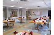 A spacious dining room with multiple square wooden tables, each set with folded red napkins and small floral centerpieces. The room has a tiled floor with a pattern of beige and blue squares, fluorescent ceiling lights, and large windows with valance curtains. There is a bulletin board and some furniture along the far wall.