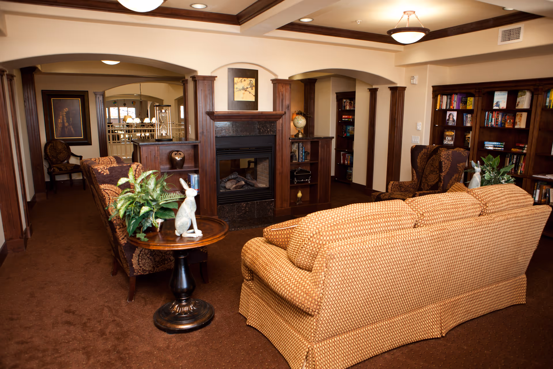 A cozy living room area in a retirement facility featuring a patterned sofa and two armchairs arranged around a wooden fireplace with built-in shelves. The room has warm brown carpeting, wooden trim, and bookshelves filled with books. A small round table with a plant and a decorative rabbit figurine is placed beside the sofa. The background shows an adjacent dining area with a chandelier.