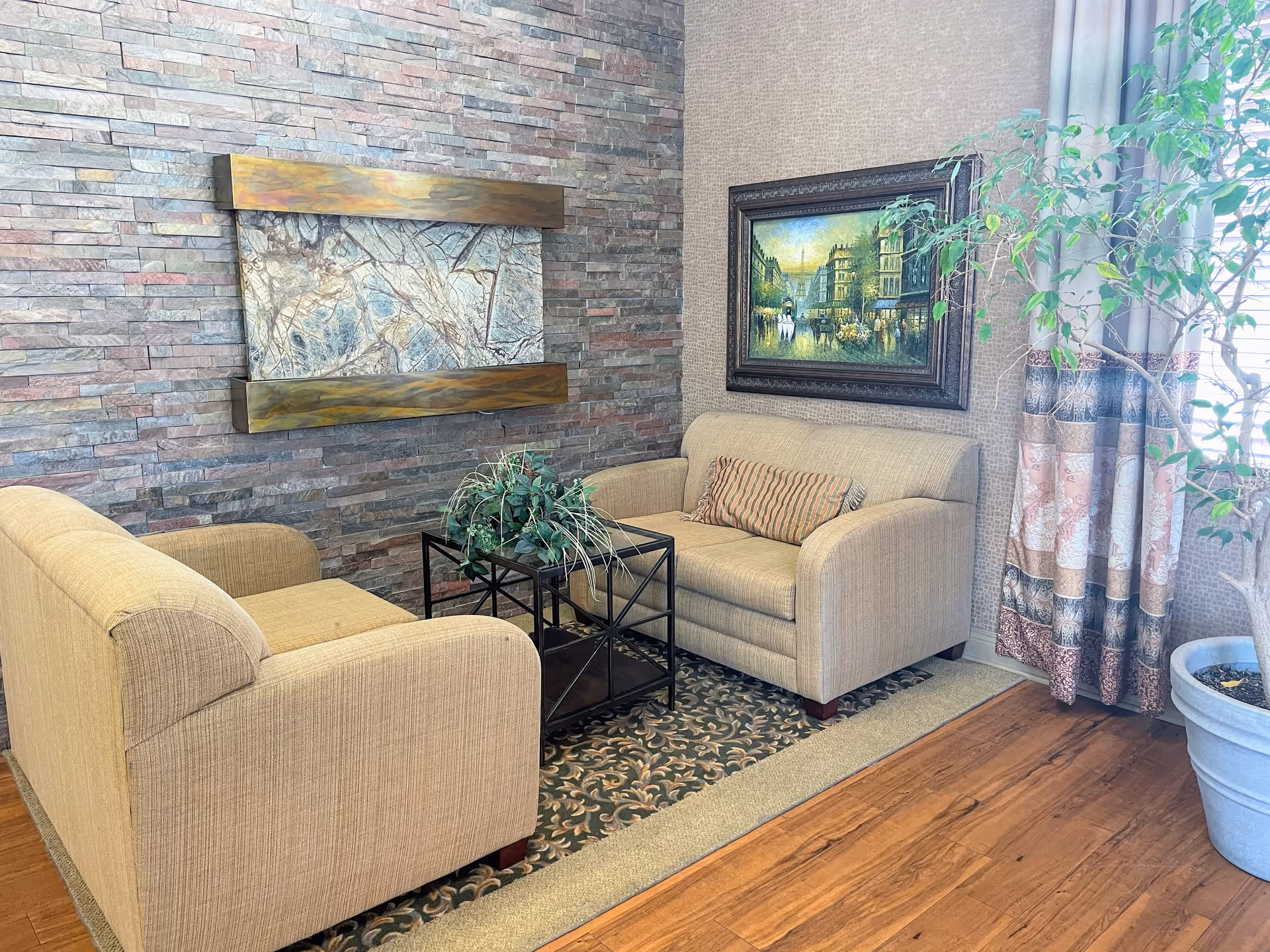 Cozy lounge area with two beige sofas facing a glass-top coffee table, a stone accent wall, framed artwork, and a potted plant by a curtained window.
