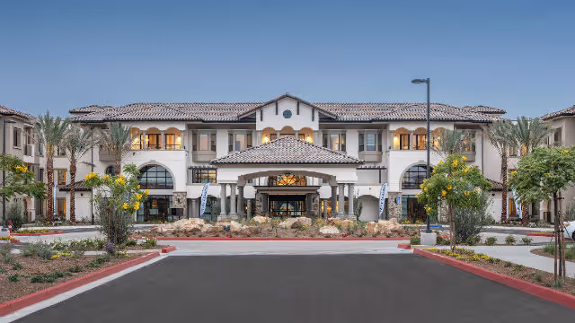 Front exterior view of Silvergate Rancho Bernardo, a two-story building with a tiled roof, arched windows, and a covered entrance. The driveway is lined with palm trees and landscaped plants under a clear blue sky.