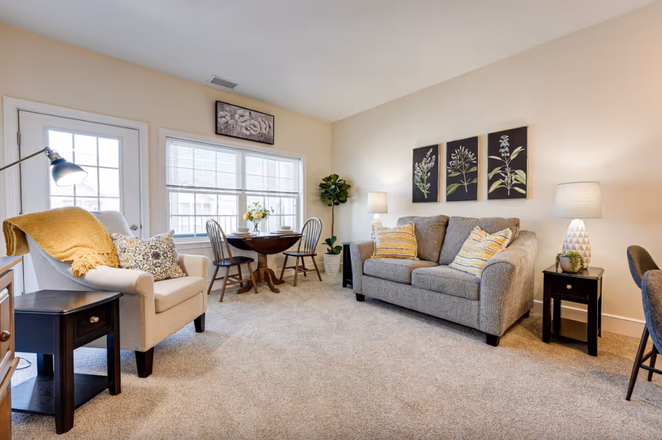 Cozy living room with a gray sofa, upholstered armchair, round dining table by a large window, lamps, and botanical wall art.