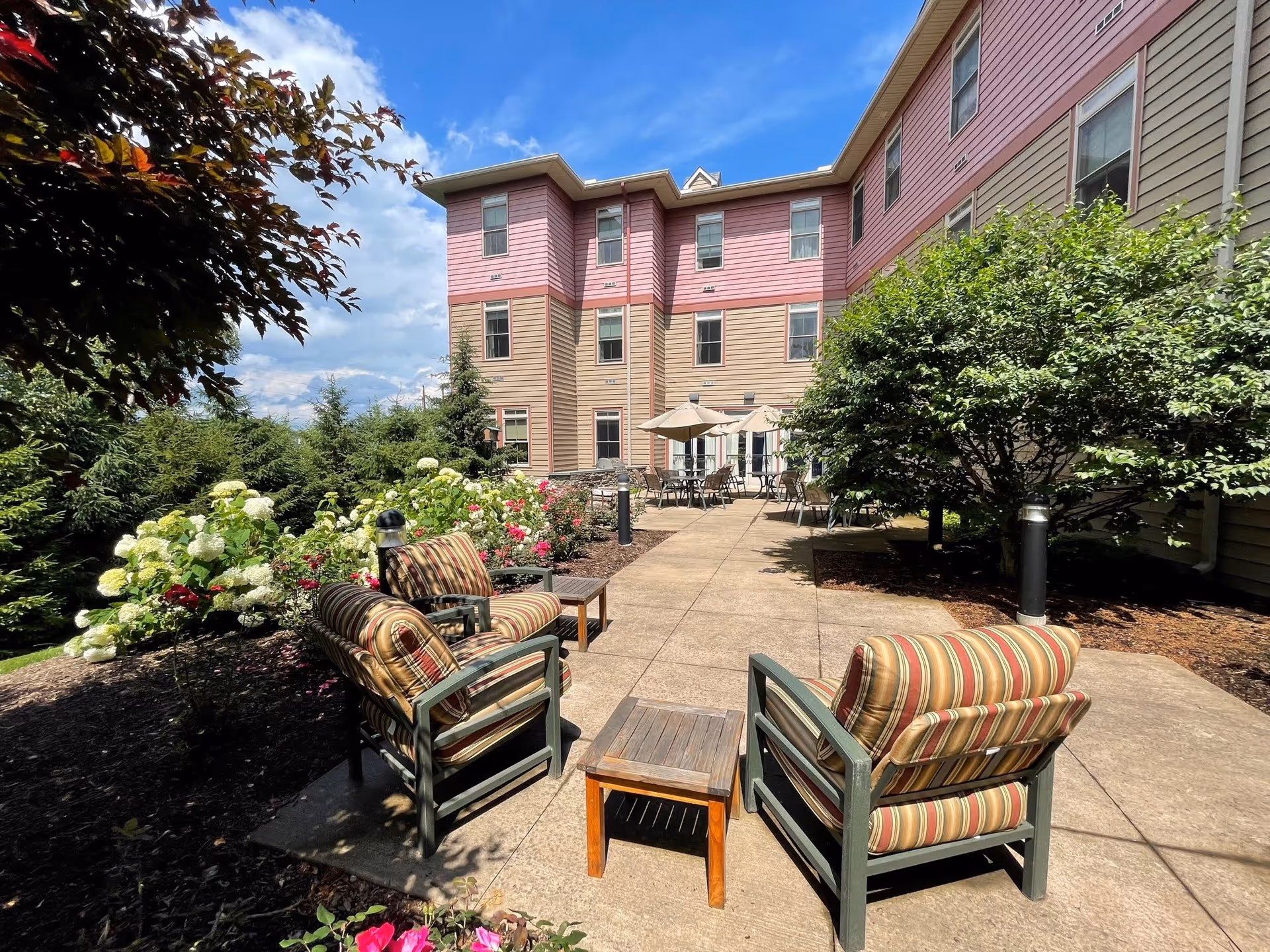 Outdoor patio area at The Pines Senior Living with cushioned chairs and small tables on a paved walkway, surrounded by flowering bushes and greenery, under a partly cloudy blue sky.