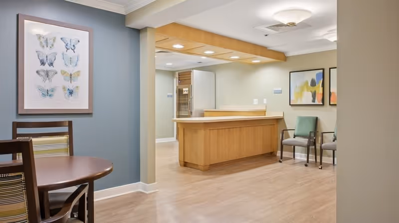 Interior view of a senior living community common area featuring a wooden reception desk with overhead lighting, two chairs against the wall with abstract artwork above them, and a round wooden table with chairs in the foreground. The walls are painted in soft neutral tones with one accent wall in blue displaying framed butterfly artwork.