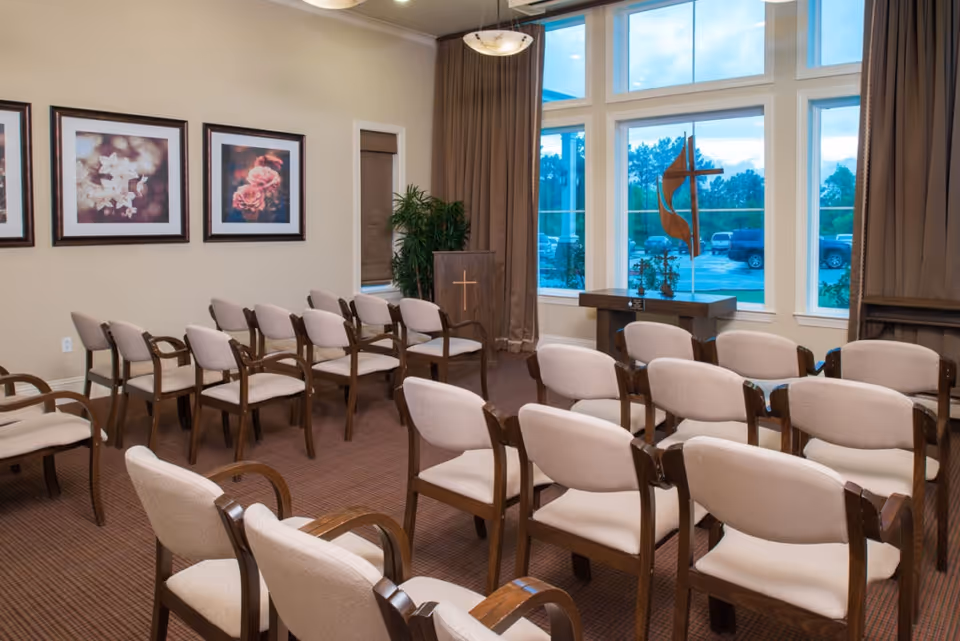 A small chapel or meeting room with rows of beige cushioned chairs with wooden frames arranged facing a wooden podium with a cross. Large windows behind the podium show a parking lot outside. The walls have framed floral artwork, and there are brown curtains on the windows.