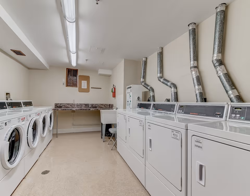 Laundry room with multiple front-loading washing machines on the left and top-loading dryers on the right. There is a folding counter at the back with a chair and a utility sink. The walls are light-colored and there are exposed ventilation pipes above the dryers.
