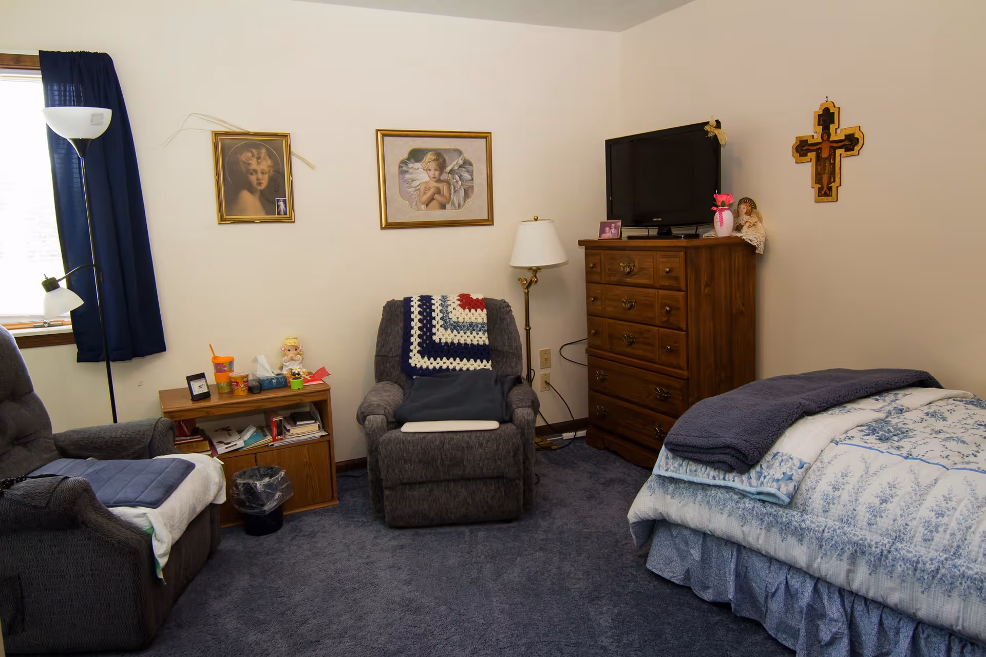 A cozy bedroom with a bed covered in blue and white bedding, a wooden dresser with a TV on top, two comfortable armchairs, a small wooden side table with various items, a floor lamp, and framed pictures on the wall including a cross.