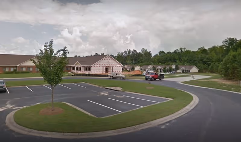 View of a senior living facility parking lot with a few cars parked and a building under construction in the background, surrounded by trees and greenery under a cloudy sky.