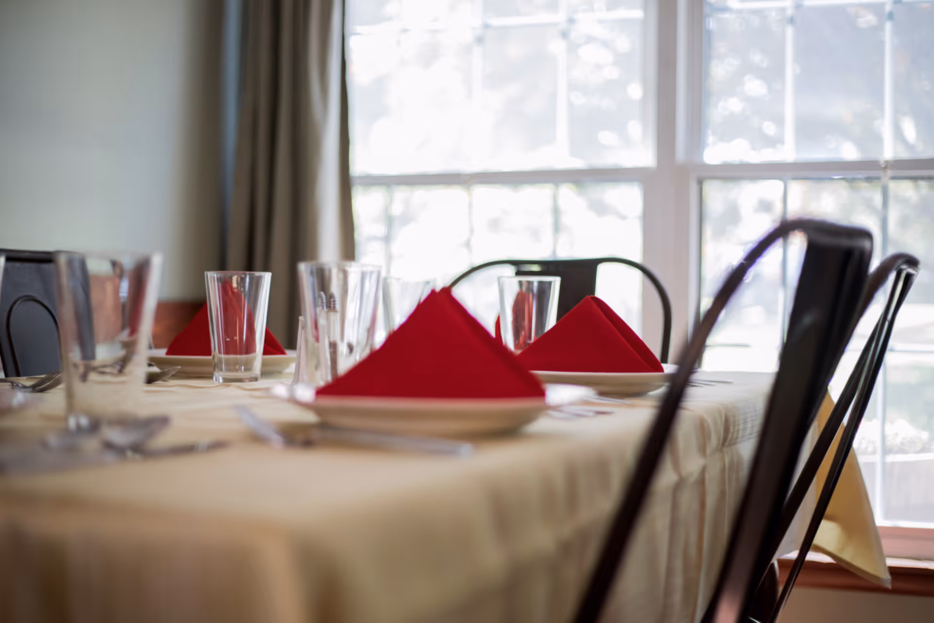 A dining table set with clear drinking glasses, white plates, silverware, and red folded napkins in front of a large window with curtains.