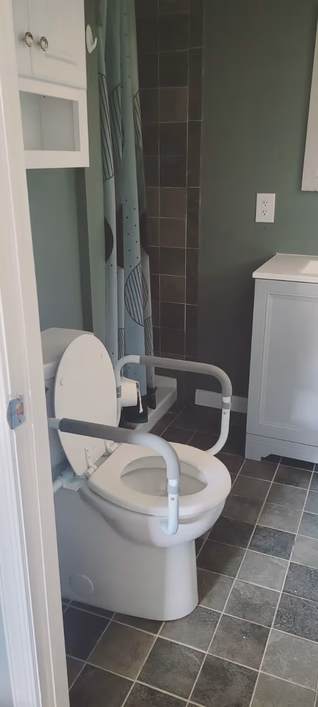 A bathroom with a white toilet equipped with safety rails on both sides. The floor is tiled with square tiles in shades of gray. There is a shower area with a curtain featuring a geometric pattern, and a white vanity with a sink and a power outlet on the wall above it. The walls are painted a muted green color.
