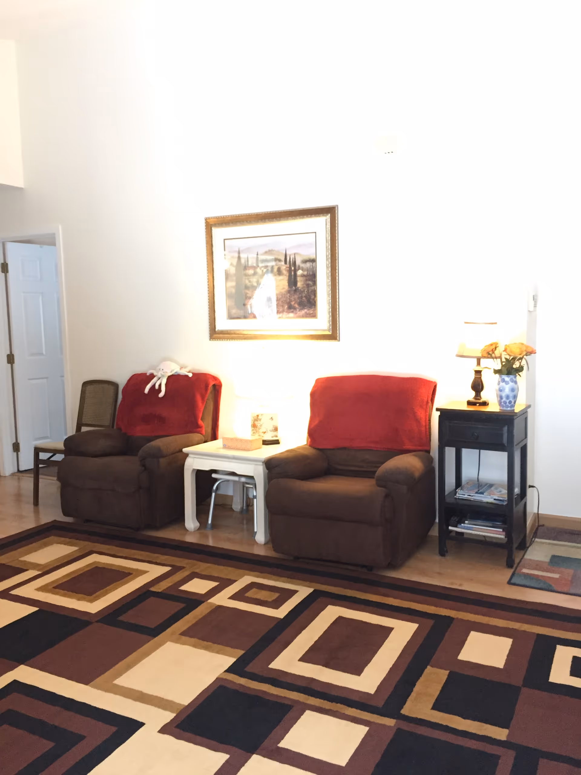 A cozy living room area with two brown recliner chairs each covered with a red throw blanket. Between the chairs is a small white side table with a lamp and a tissue box. To the right of the right chair is a black side table with a blue vase holding orange flowers and a small lamp. A framed landscape painting hangs on the white wall above the chairs. The floor features a patterned area rug with geometric shapes in shades of brown, beige, and black.