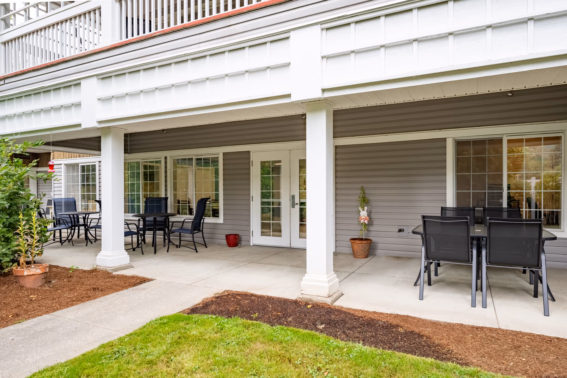 Covered outdoor patio area at Birchview Memory Care with multiple black metal tables and chairs arranged on a concrete floor. The patio is supported by white columns and has a gray exterior wall with windows and a glass door. There are potted plants and a small patch of grass and mulch in the foreground.