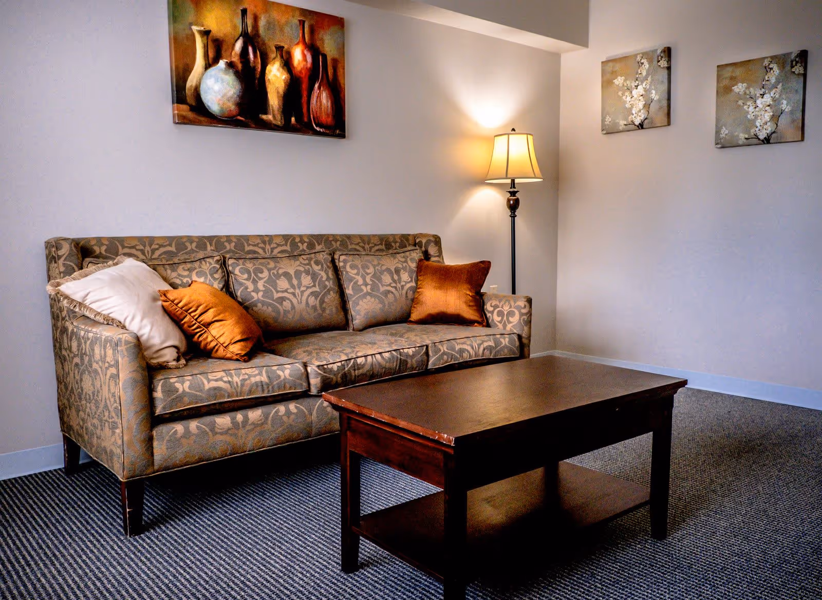 A cozy living room area with a patterned sofa adorned with three cushions, a wooden coffee table in front, a floor lamp emitting warm light in the corner, and three paintings on the walls featuring vases and floral designs.