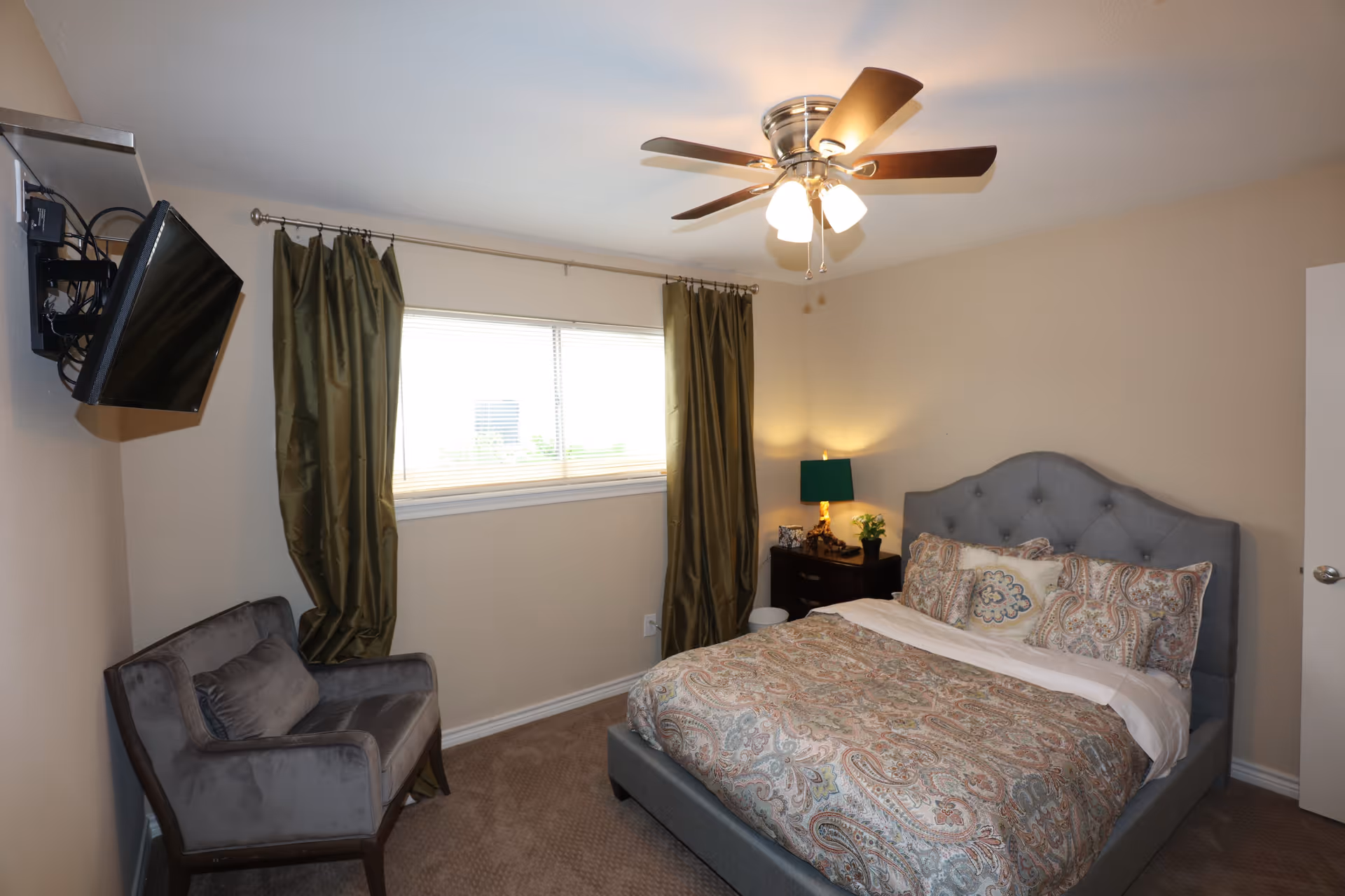 A cozy bedroom with a gray upholstered bed featuring a paisley patterned comforter and multiple pillows. Next to the bed is a dark wooden nightstand with a green lamp, a small plant, and a photo frame. A gray armchair is positioned near a window with green curtains. A ceiling fan with lights is mounted on the ceiling, and a wall-mounted TV is angled towards the bed.