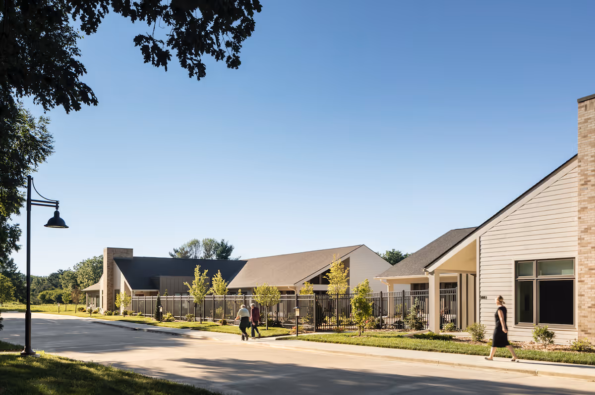 Exterior view of a single-story senior living facility with landscaped grounds, a streetlamp, and people walking along the sidewalk.