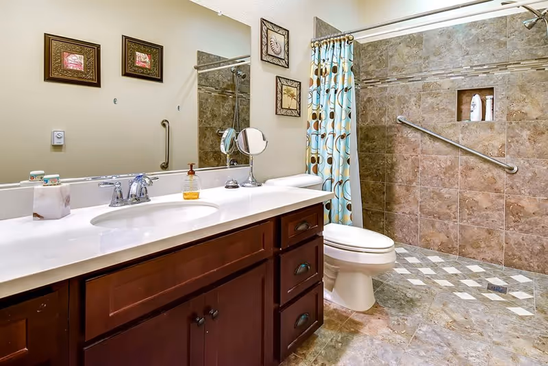Bathroom with a sink vanity and mirror, toilet, and a tiled walk-in shower with a grab bar and patterned shower curtain.