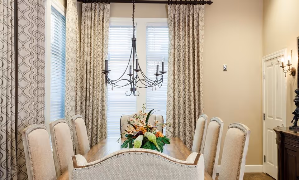 A well-lit dining room with a wooden dining table surrounded by eight upholstered chairs. A decorative floral centerpiece is placed on the table. The room features large windows with patterned curtains and a black wrought-iron chandelier hanging above the table. Beige walls and a wooden sideboard with a sculpture are visible.