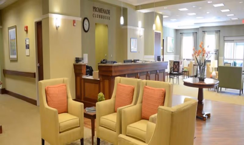 Interior view of a senior living facility lounge area with four beige armchairs featuring orange cushions arranged around a small wooden table. In the background, there is a reception desk with a sign above it reading 'PROMENADE CLUBHOUSE'. The room is well-lit with ceiling lights and wall sconces, and there are large windows with curtains letting in natural light. Additional seating and tables are visible further back in the room.