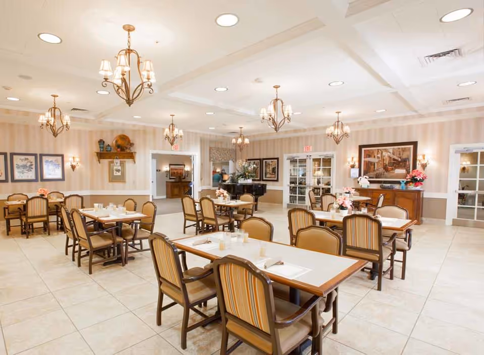 A spacious dining room with several tables and chairs arranged neatly. The room features beige tiled floors, striped wallpaper, and multiple chandeliers hanging from a white coffered ceiling. The tables are set with placemats, napkins, and glasses. The walls are decorated with framed artwork and floral arrangements. There is a piano in the background and double glass doors leading to other rooms.