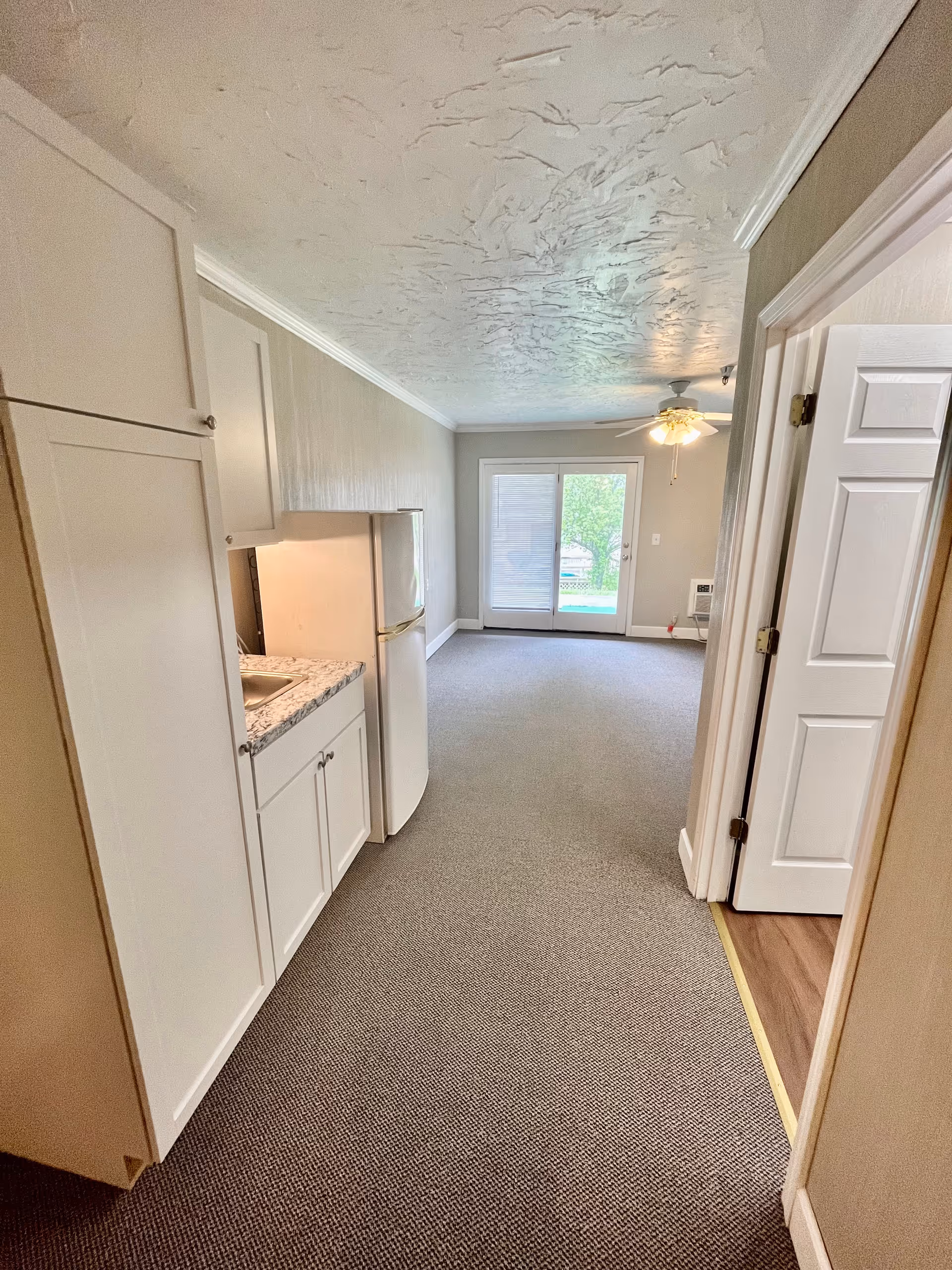 Interior view of a small apartment or living space with a kitchenette on the left featuring white cabinets and a countertop with a sink. A refrigerator is next to the kitchenette. The room has carpeted flooring and leads to a living area with a ceiling fan and glass sliding doors that open to an outdoor area. A doorway on the right leads to another room with wooden flooring.