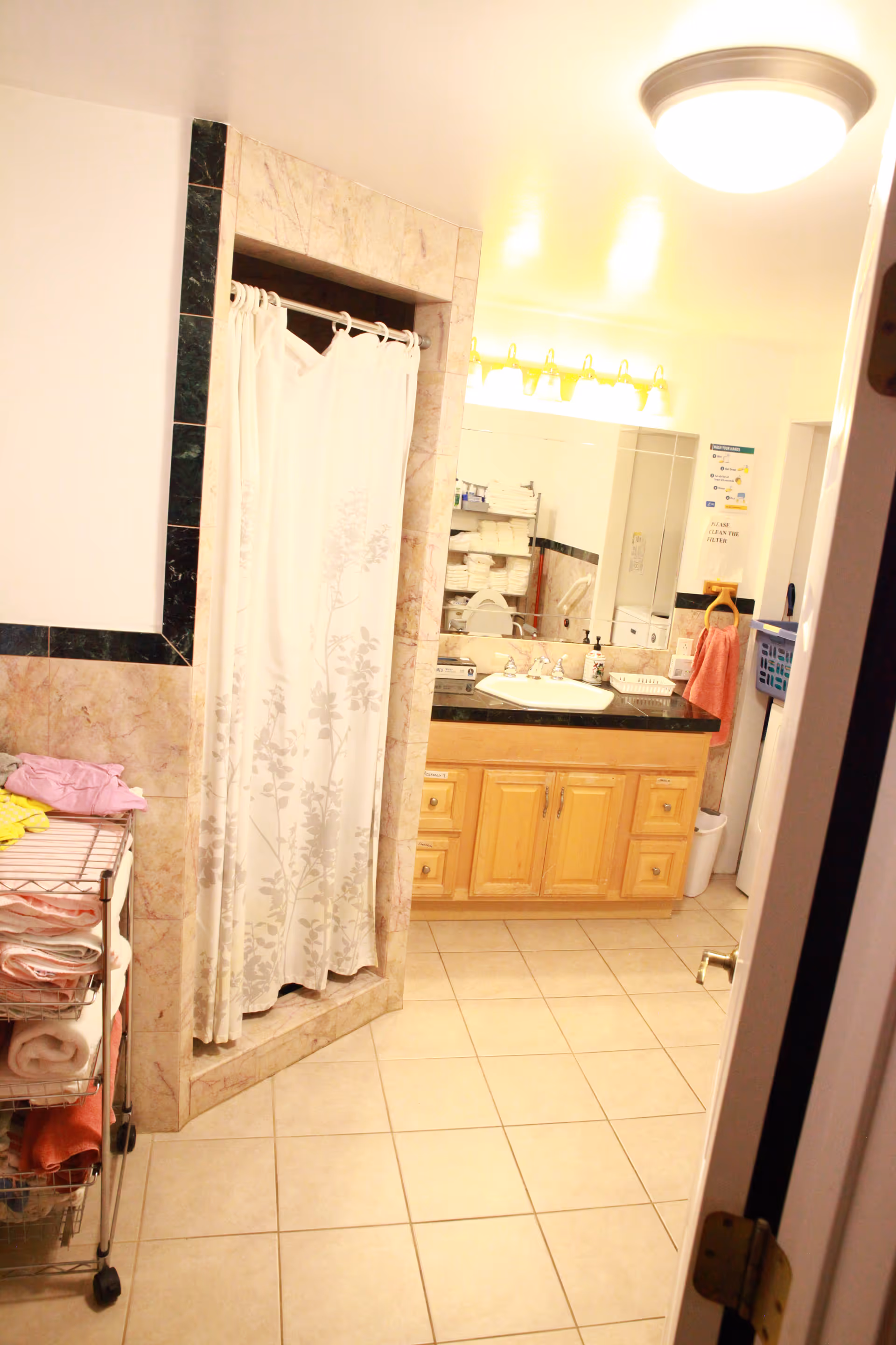 A bathroom with beige tiled floor and walls, featuring a shower with a white floral shower curtain, a wooden vanity with a black countertop, a sink, and a large mirror above it. There is a metal rack with folded towels and clothes on the left side, and a ceiling light fixture illuminating the room.