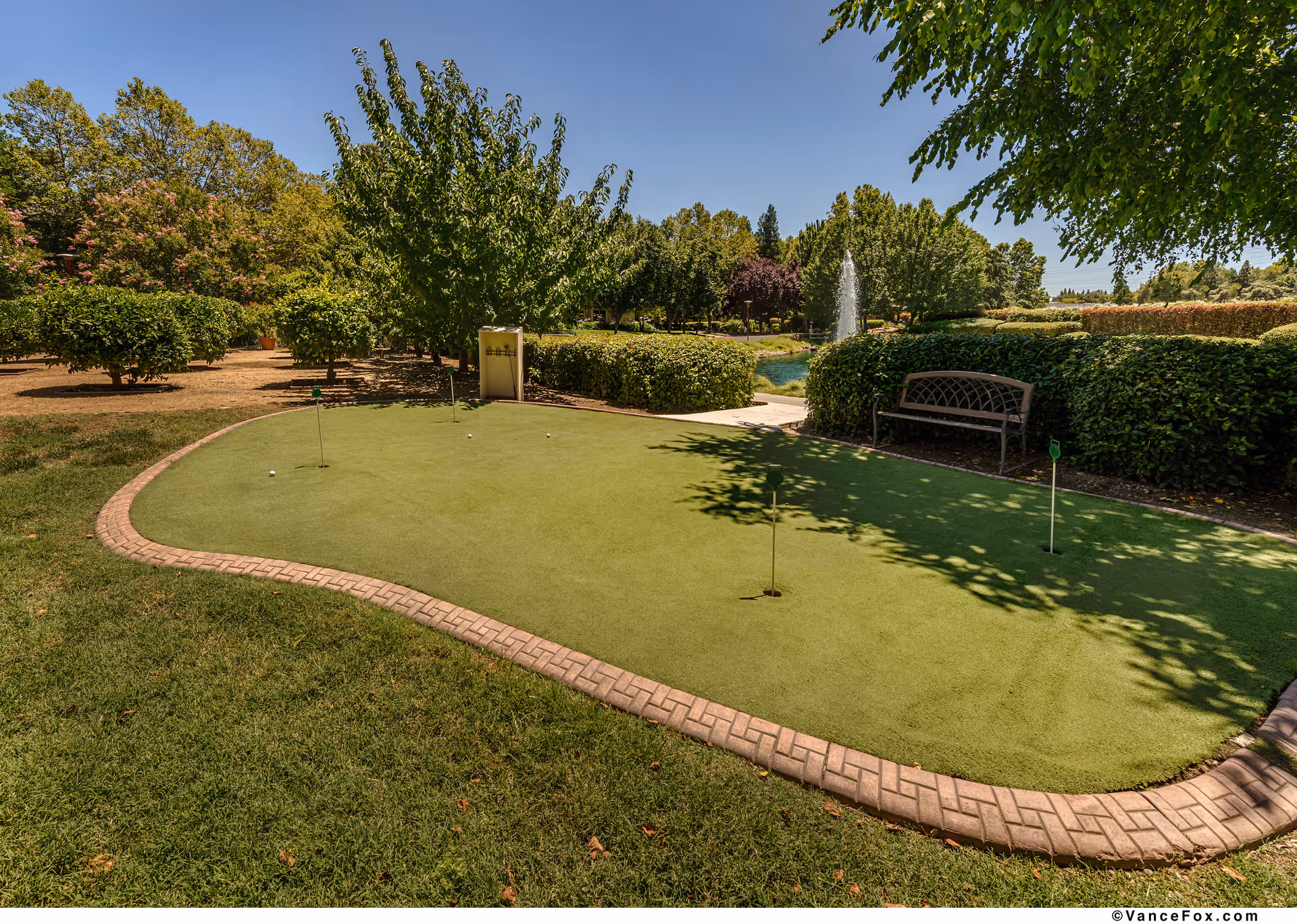 A small outdoor putting green with several golf holes and flags, surrounded by green grass, bushes, and trees. There is a bench on the right side and a water fountain in the background under a clear blue sky.