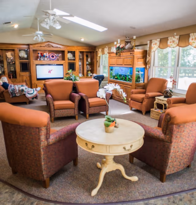 A cozy living room with multiple upholstered armchairs arranged around a round wooden table with a small potted plant. The room features a large wooden entertainment center with a TV, decorative items, and a large aquarium. Large windows with floral valances allow natural light to fill the space.