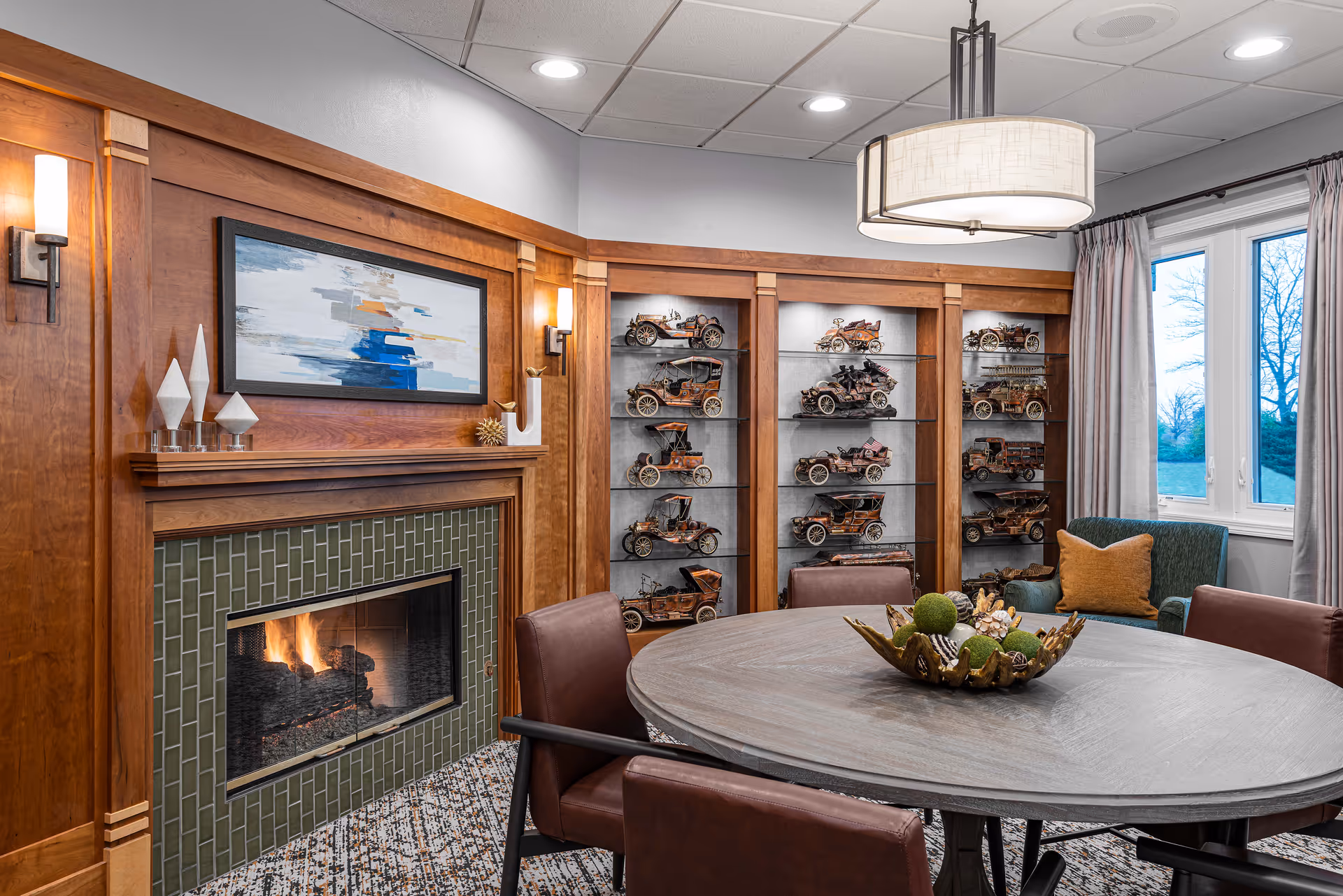 A cozy interior room with a round wooden table surrounded by brown leather chairs. A decorative bowl with green and natural elements sits on the table. Behind the table is a fireplace with a green tiled surround and a wooden mantel, above which hangs an abstract painting. To the right of the fireplace are built-in wooden shelves displaying vintage model cars. A large window with curtains lets in natural light, and a modern ceiling light fixture hangs above the table.