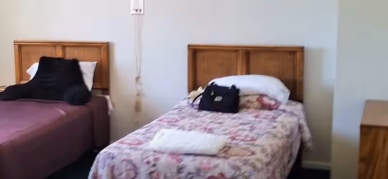 Two single beds with wooden headboards in a simple assisted living bedroom, one bed covered in a floral bedspread with a small bag on top.