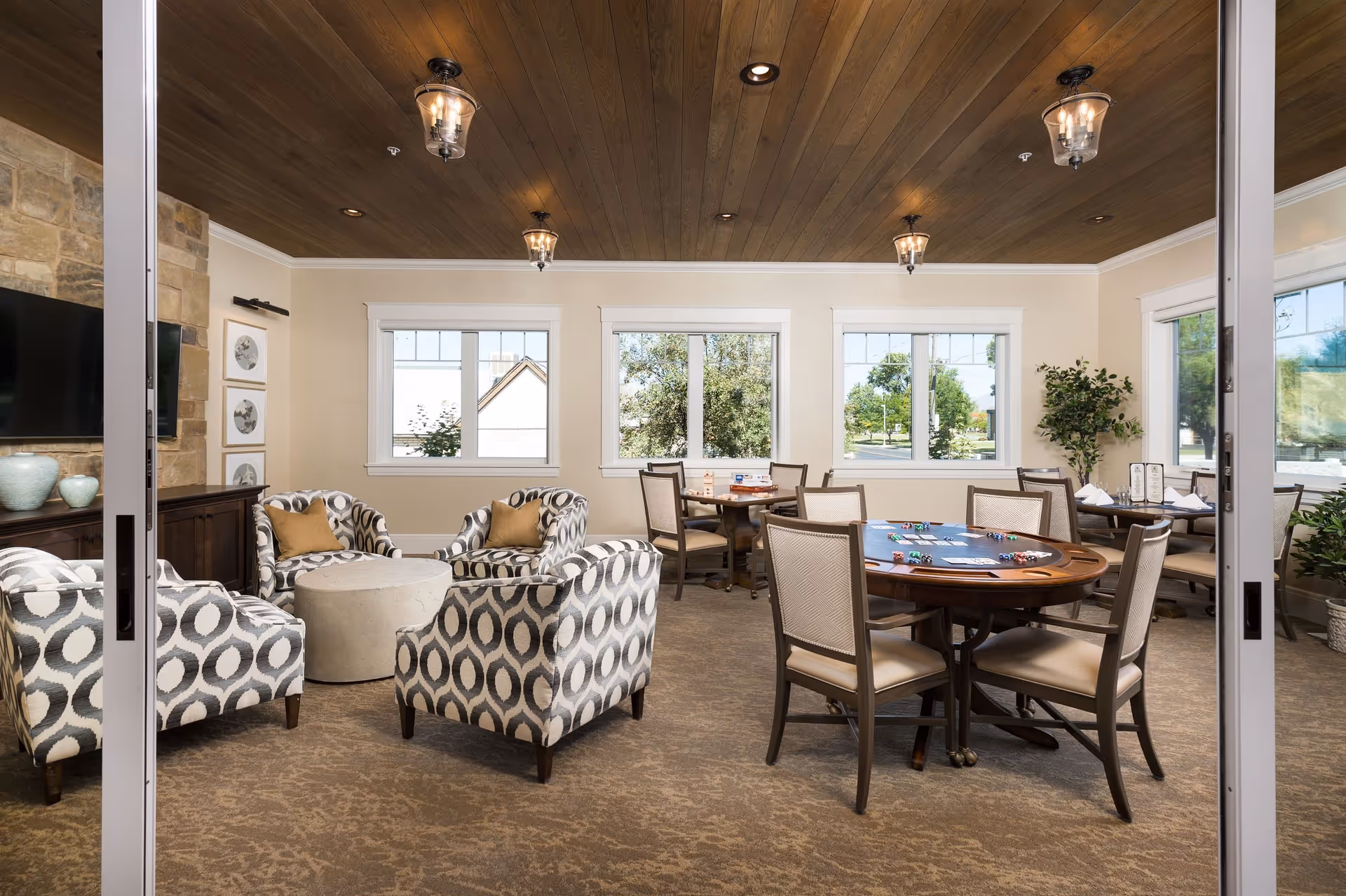 A cozy senior living common area with patterned armchairs arranged around a round ottoman, a stone fireplace with a mounted TV, and several tables with chairs set up for games and dining. Large windows let in natural light and show greenery outside. The ceiling is wooden with hanging lantern-style lights.