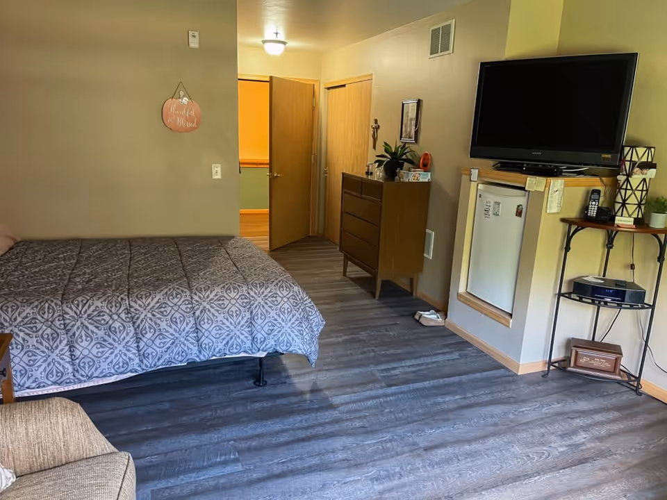 A furnished studio bedroom with a bed, dresser, wall-mounted TV, mini-fridge and chair on wood-look flooring.