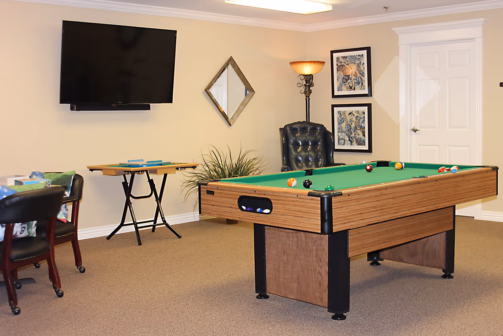 A recreational room with a pool table in the center, a black leather chair, a standing lamp, a wall-mounted flat screen TV, a small table with board games, and framed artwork on the wall.
