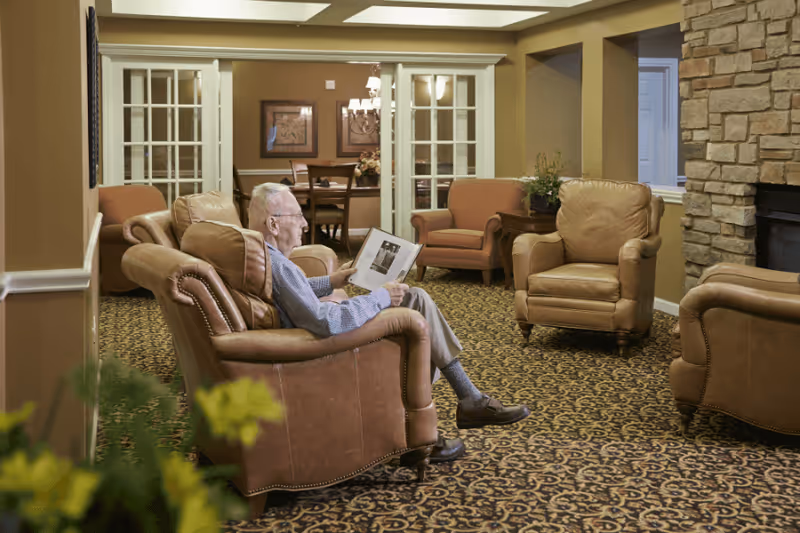 An elderly man sitting in a brown leather armchair in a cozy living room area, reading a book or magazine. The room features patterned carpet, additional armchairs, a stone fireplace on the right, and a dining area visible through glass-paneled double doors in the background.