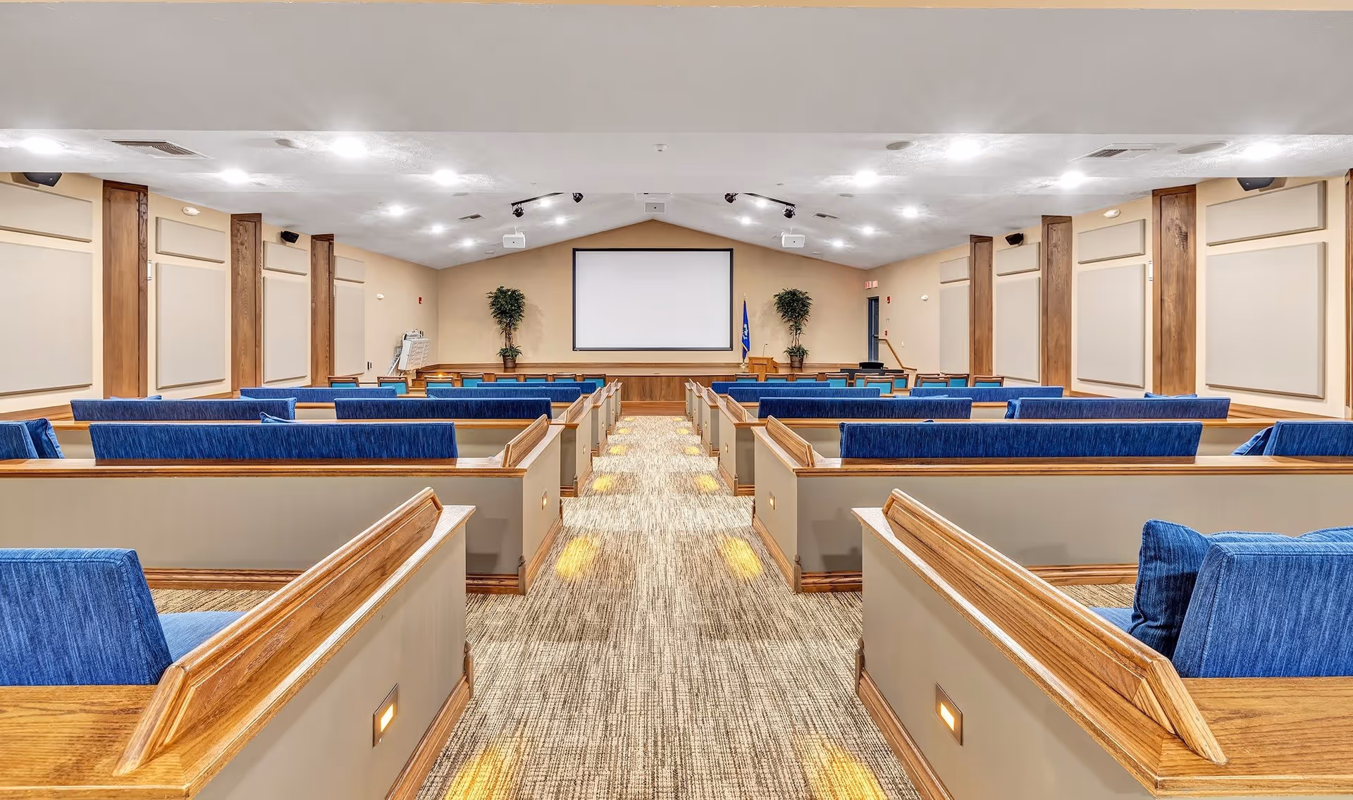 Interior view of a large meeting or presentation room with rows of wooden pew-style seating with blue cushions, a carpeted aisle with floor lighting, beige walls with wooden accents, and a large blank projection screen at the front flanked by two potted plants and a podium.