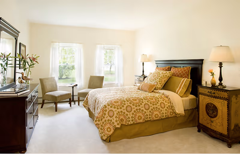 A bright and cozy bedroom with a large bed featuring patterned bedding and multiple pillows. There are two beige chairs and a small round table near two windows with sheer white curtains. The room includes a dark wooden dresser with a mirror and decorative items, as well as two bedside tables with lamps and a small vase with flowers.