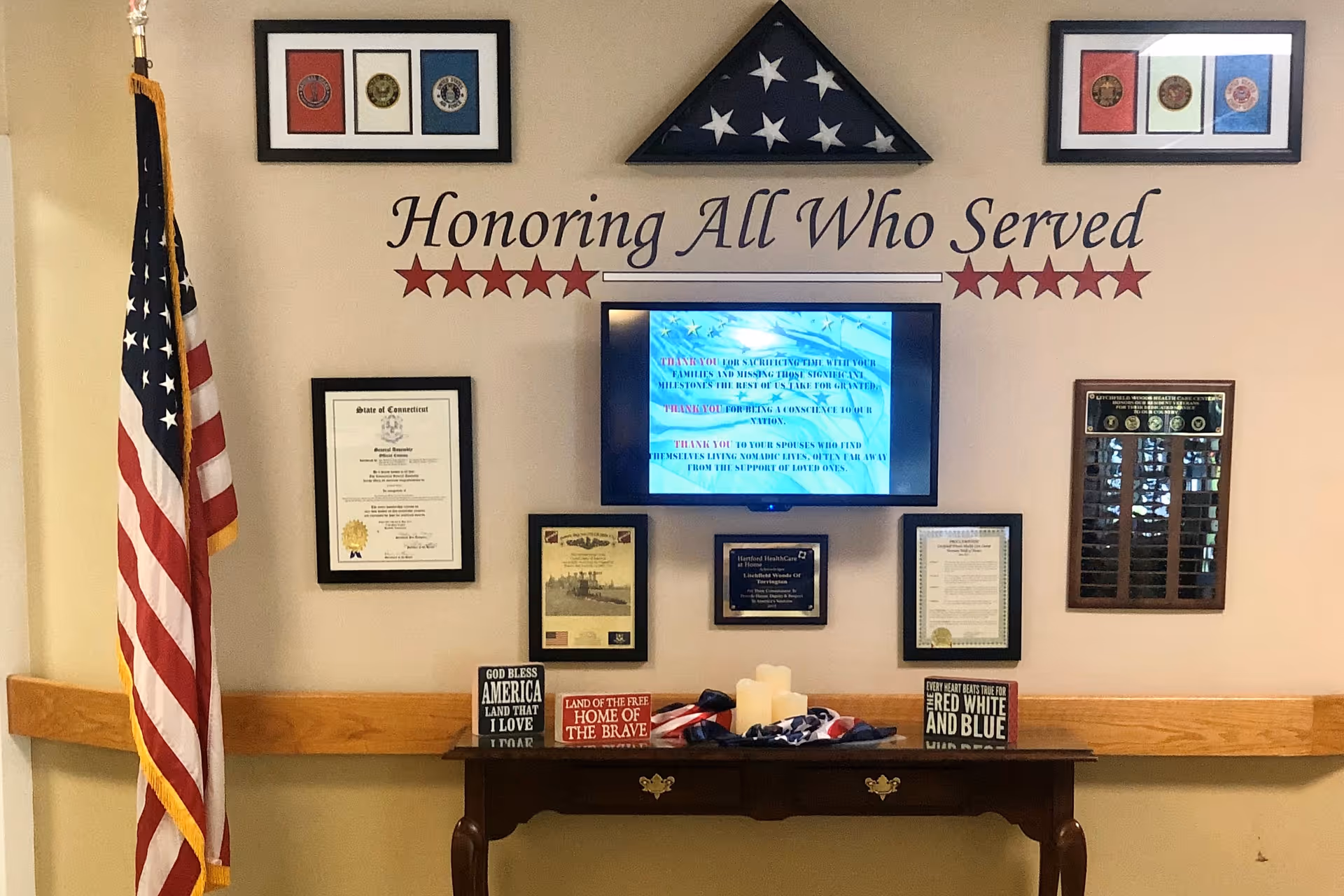 A memorial wall display honoring military service members with the text 'Honoring All Who Served' above a folded American flag in a triangular case. The wall features framed certificates, plaques, and patriotic decorations including an American flag on a stand to the left and small signs with patriotic messages on a wooden table below the display.