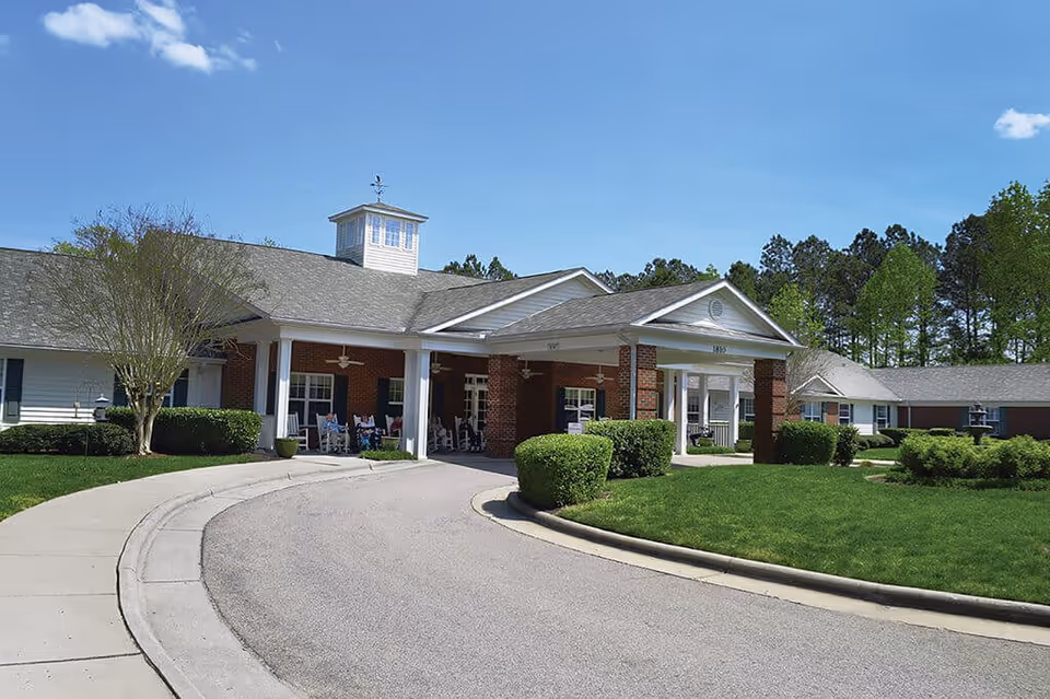 Exterior view of Spring Arbor of Raleigh, showing a single-story building with a covered entrance supported by white columns. The building has a brick facade with white siding and a cupola on the roof. There is a curved driveway leading to the entrance, surrounded by neatly trimmed bushes and green grass under a clear blue sky.