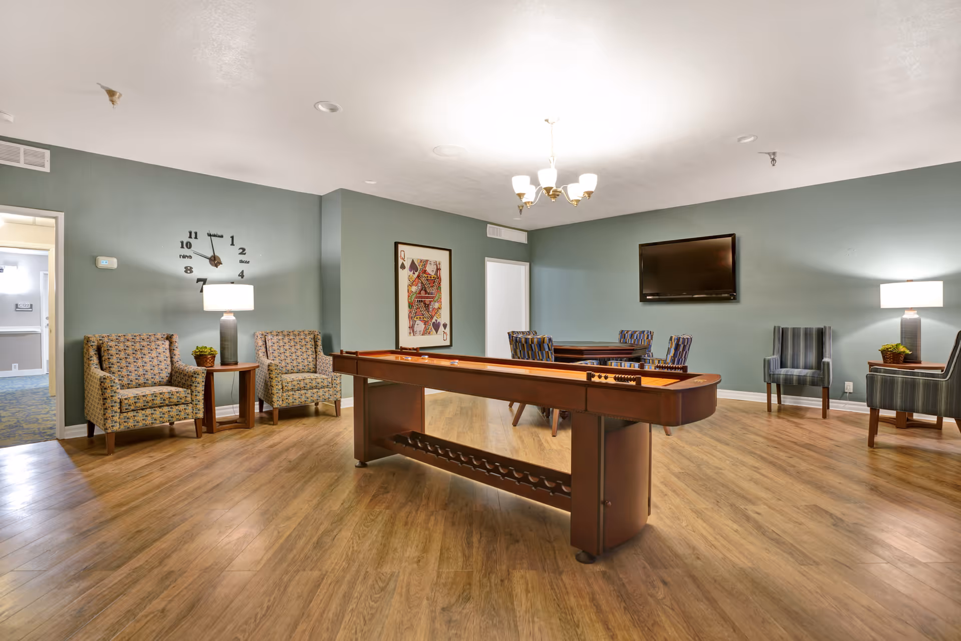 A spacious game room with a shuffleboard table in the center, surrounded by several upholstered chairs and small tables with lamps and plants. The walls are painted green, and there is a large wall clock and a framed playing card artwork. A flat-screen TV is mounted on the wall, and the floor is wood.