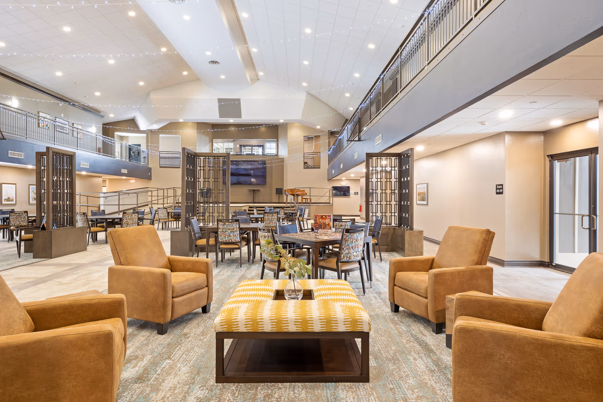Spacious senior living facility common area with high ceilings, multiple seating arrangements including four brown armchairs around a patterned ottoman, and several dining tables with chairs. The room features a second-floor balcony, modern lighting, and neutral-toned walls and flooring.
